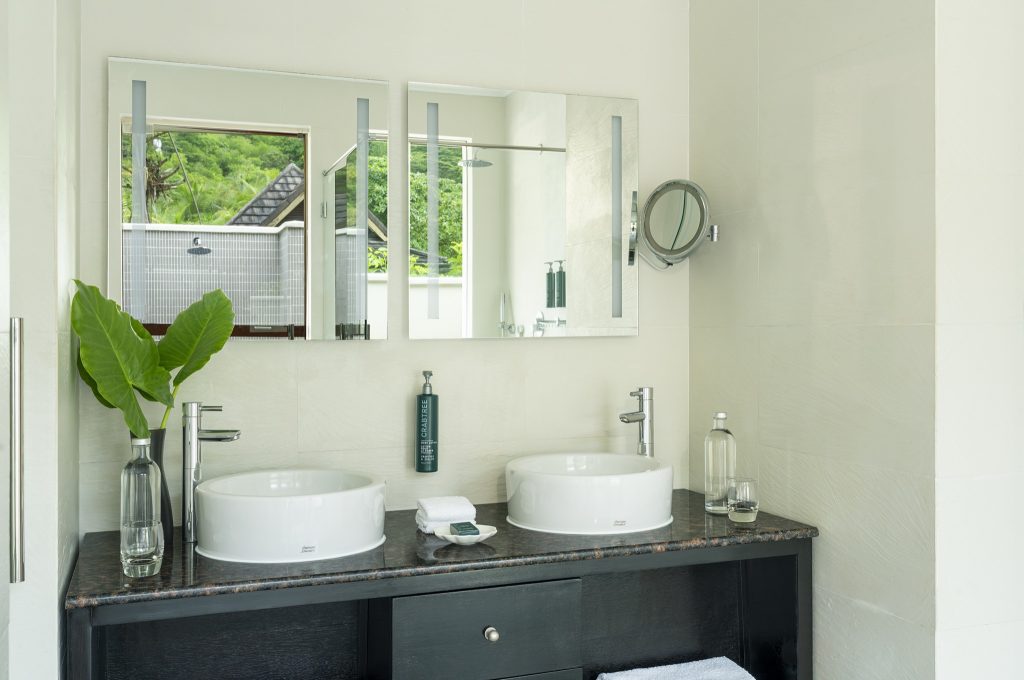 Modernes Badezimmer der Garden Villa im NIVA Labriz Seychelles mit zwei weißen Keramikwaschbecken auf dunkler Steinplatte