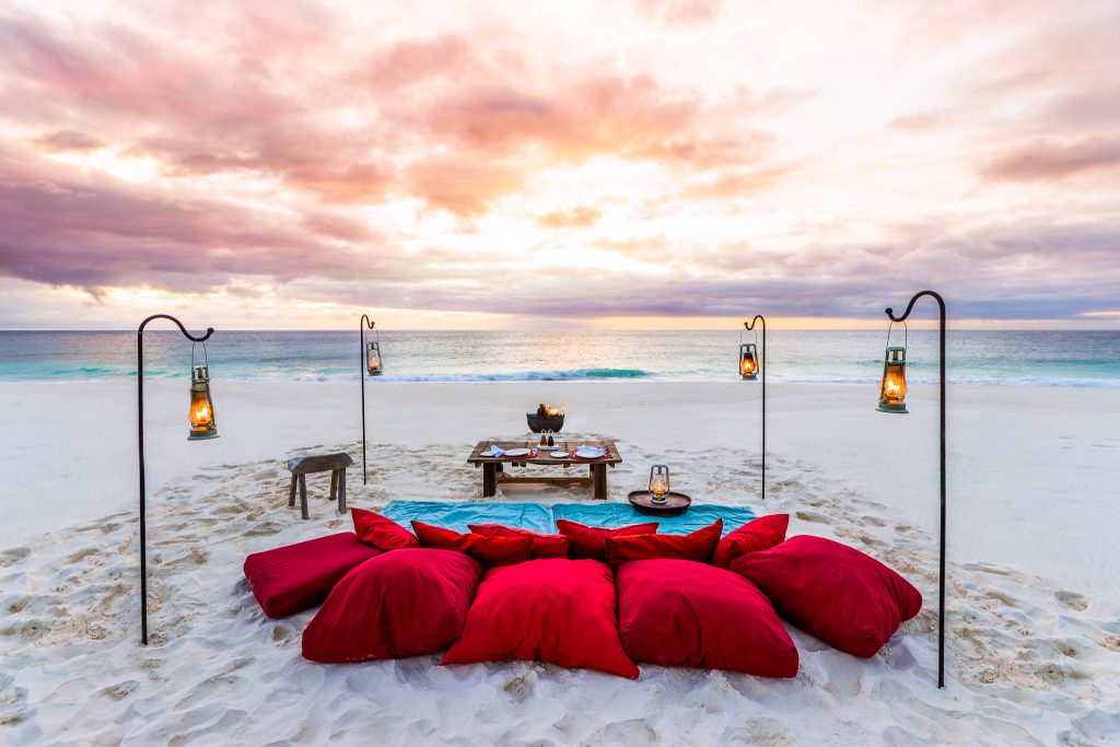 Strand mit roten Kissen, blauer Decke und Holztisch, Laternen und Meer im Hintergrund bei Sonnenuntergang