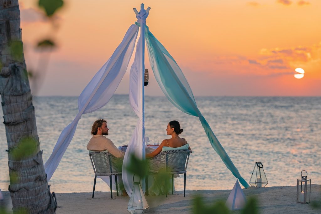 Strand-Dinner bei Sonnenuntergang mit zwei Stühlen aus Netzmaterial unter Baldachin aus weißen und aquafarbenen Stoffen