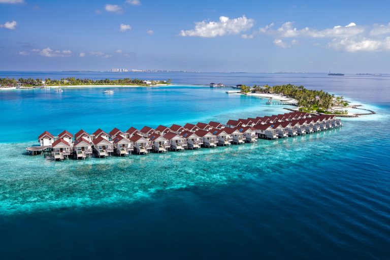 Reihe von hölzernen Wasserbungalows auf Stelzen im türkisblauen Meer mit grüner Insel im Hintergrund