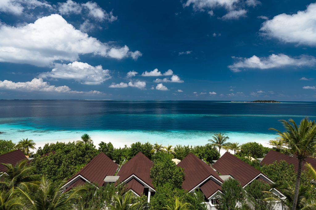 Luftaufnahme von OBLU SELECT Lobigili Sunnest Beach Pool Villa mit dunkel gedeckten Dächern, Palmen.