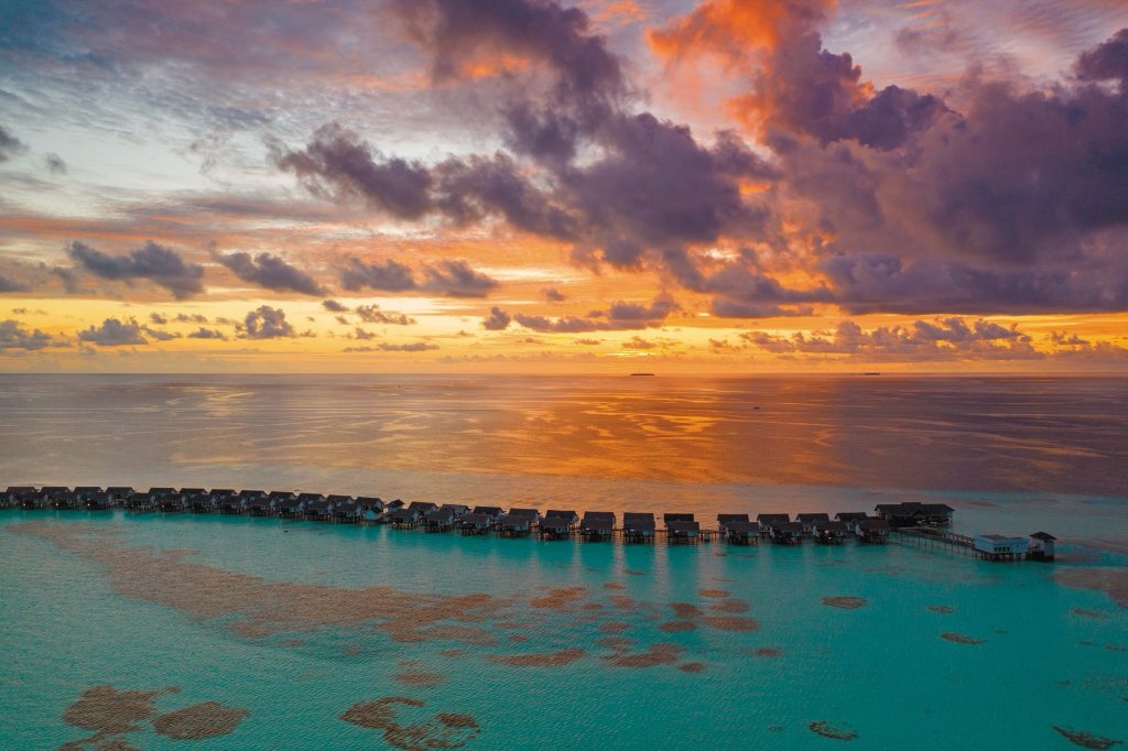 Reihe von Bungalows auf Stelzen verbunden durch Stege über türkisfarbenem Wasser bei Sonnenuntergang