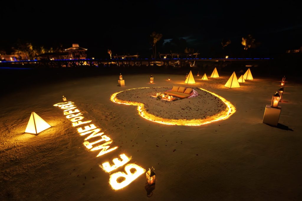 Romantische Stranddinner-Anordnung mit Kerzen, Rosenblättern und Lichtern bei Abenddämmerung am Strand von Ozen Life Maadhoo