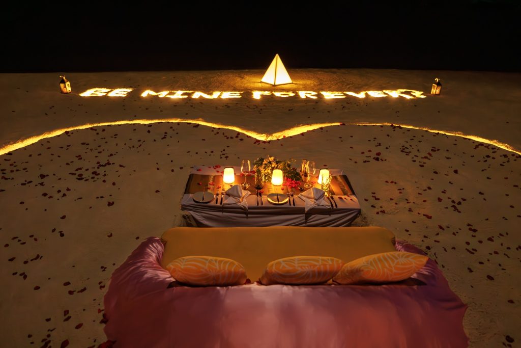 gedeckter Tisch am Strand bei Nacht mit weißen Stoffen, Kerzenlicht, Gedecken, Gläsern, Blumen und Rosenblättern