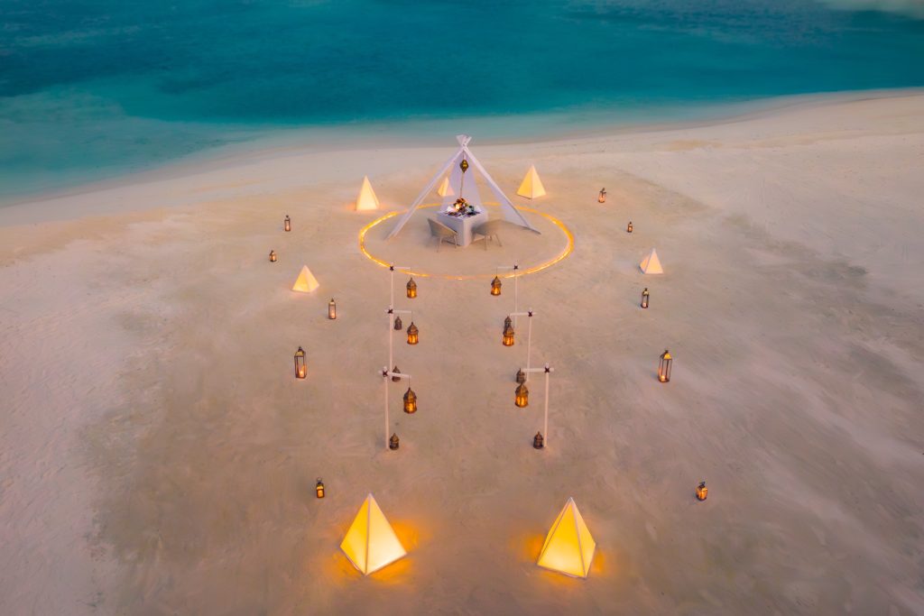 Romantischer Essbereich am Strand mit weißem Tipi-Zelt, beleuchteten Laternen und Sand bei Sonnenuntergang