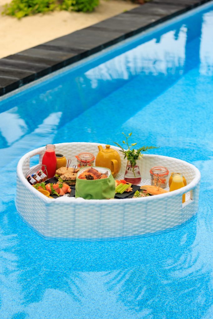 Schwimmendes Frühstückstablett mit Croissants, Obst, Saft und Teekanne auf blauem Poolwasser im Ozen Life Maadhoo