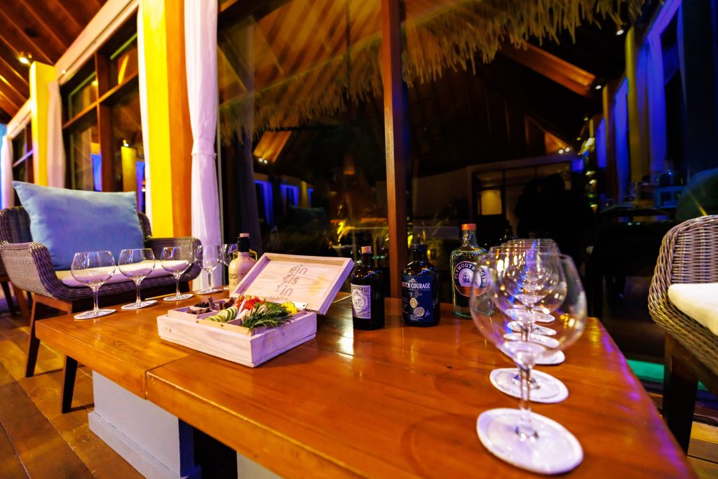 Indoor-Lounge mit Holz-Couchtisch, Gin-Flaschen, Weingläsern, Holz- und Rattanmöbeln in warmem Braun und Blau