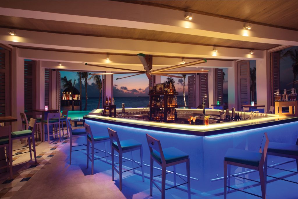 U-förmige Bar mit blau beleuchtetem Sockel, Holzbarhockern, Glasflaschen und Meerblick im Hintergrund