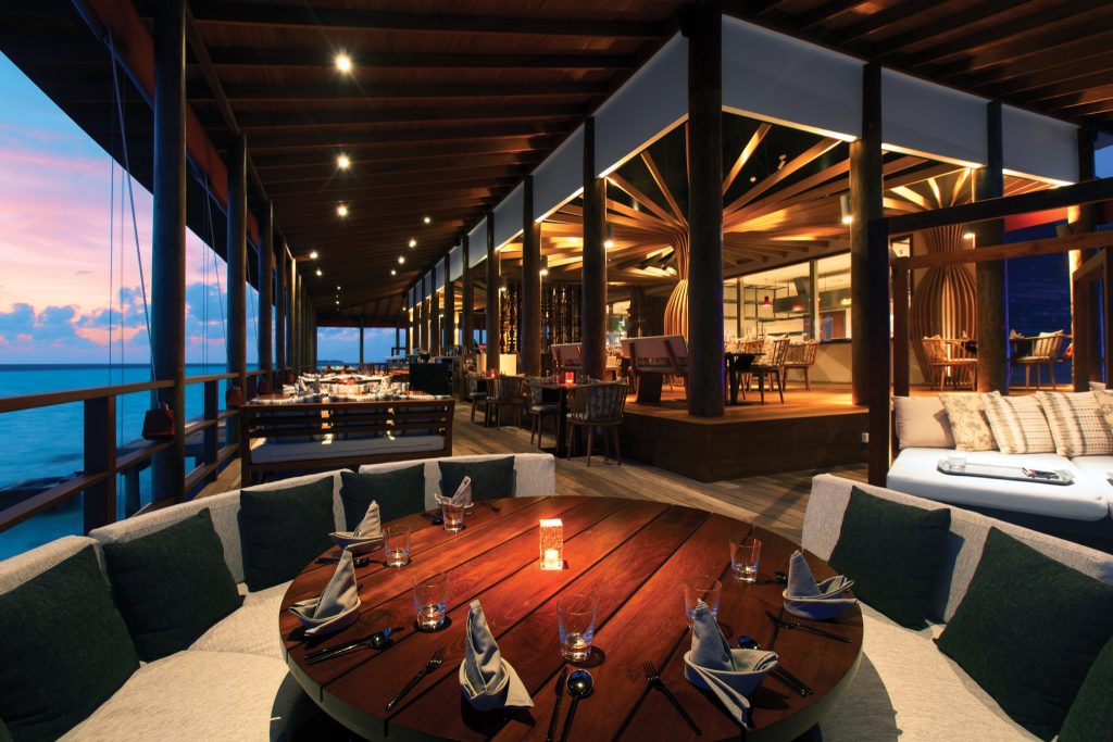 Holzterrasse des Restaurant Peking mit großer runder Tafel, Polsterbänken und Blick auf den Ozean bei Dämmerung