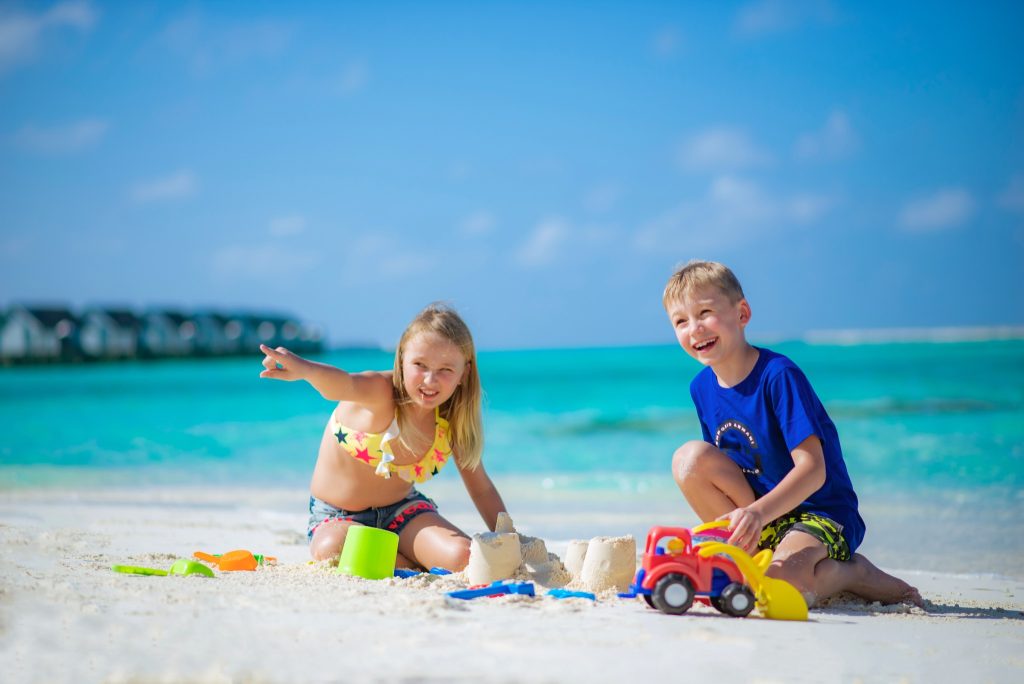 Zwei Kinder spielen mit bunten Sandspielzeugen am weißen Sandstrand vor türkisfarbenem Meer und Bungalows