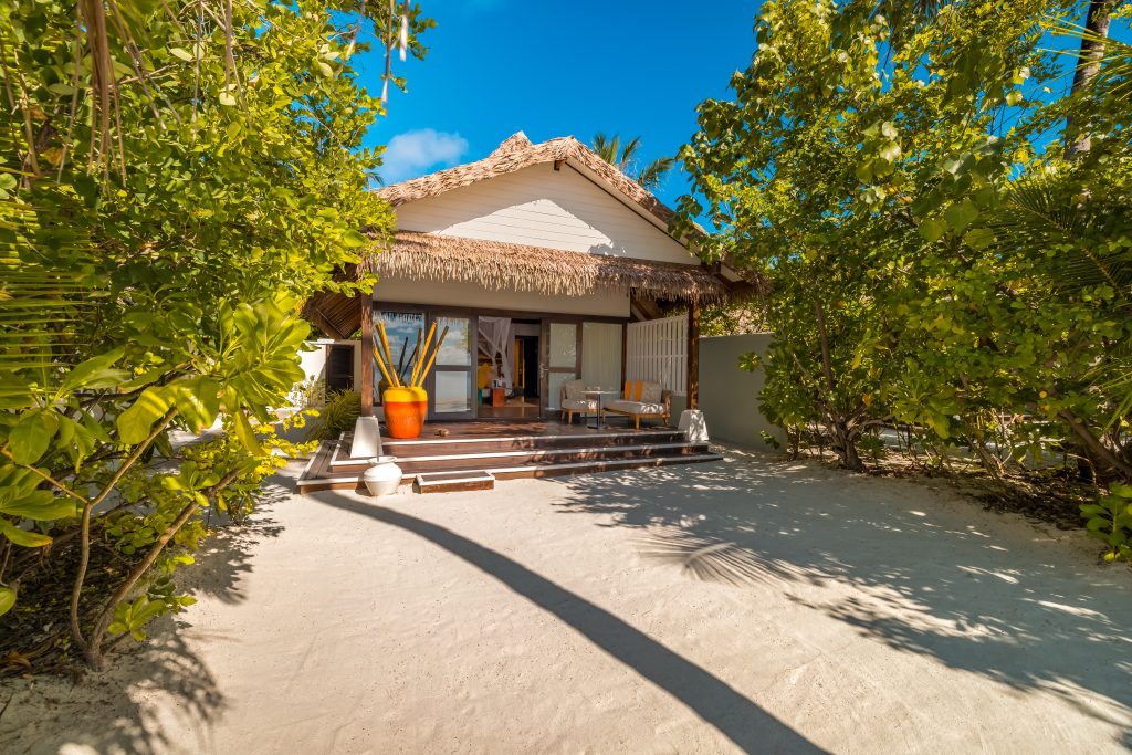 Eingang der Earth Villa im Ozen Life Maadhoo mit Holzfassade, Palmblattdach, Terrassenmöbeln und großem Blumentopf