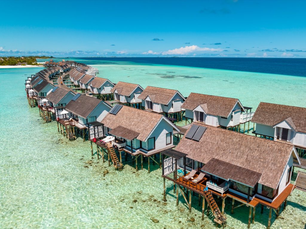 Reihe von Wind Villa Wasserbungalows auf Stelzen über türkisfarbenem Wasser verbunden durch Holzsteg bei Tageslicht