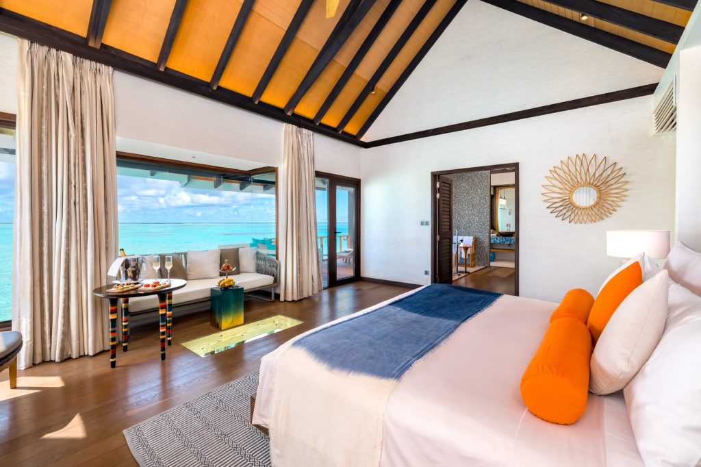 Modernes Hotelzimmer Wind Villa mit großem Bett, orangen Kissen, Holzboden und Fensterfront zum türkisfarbenen Meer
