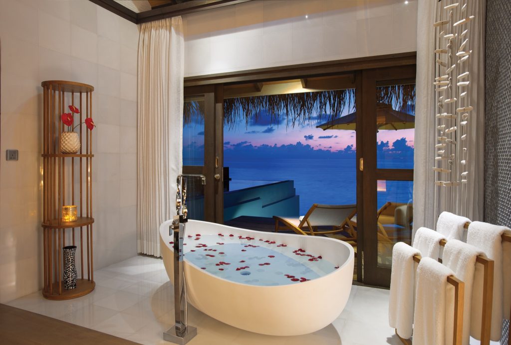 Freistehende ovale Badewanne mit Rosenblättern, weiße Handtücher auf Holzständer, Holztisch mit Vase und Kerze, Blick.