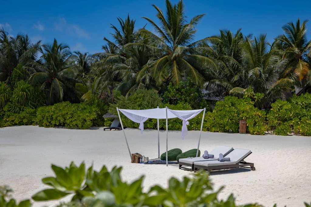 Strand mit weißem Sand, Bali-Bett mit weißem Stoffdach und zwei Holzliegen mit grauen Handtüchern