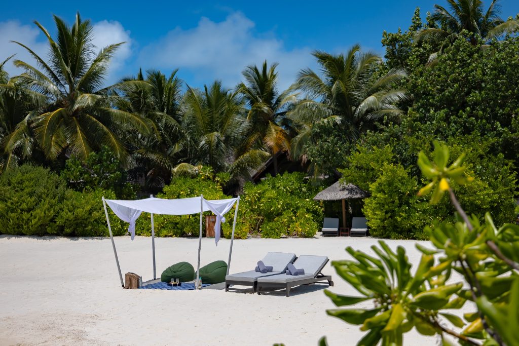 Strand mit weißem Pavillon, grünen Sitzsäcken, grauen Liegestühlen und Palmen im Hintergrund auf den