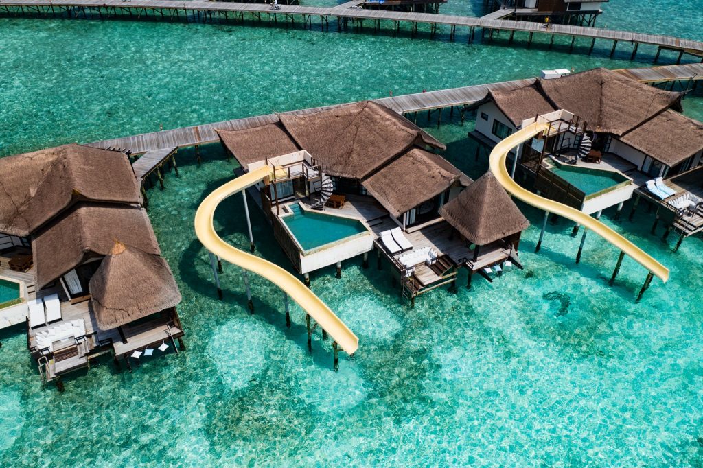 Luftaufnahme von Ocean Pool Suiten mit gelben Wasserrutschen auf Stelzen über türkisfarbenem Wasser im Ozen Reserve Bolifushi