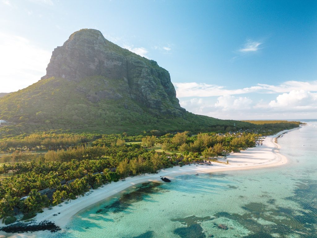 Luftaufnahme des Paradis Beachcomber Golf Resort & Spa mit weißem Sandstrand, türkisfarbenem Wasser und bewachsenem Berg