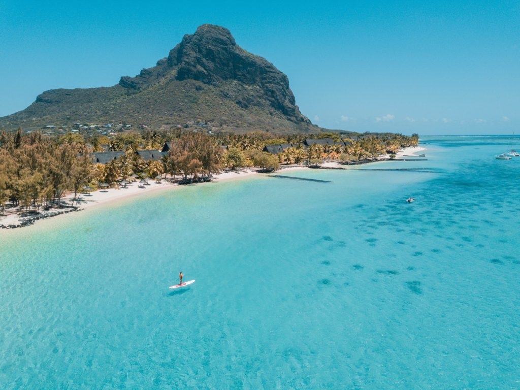 Luftaufnahme eines Strandes mit türkisblauem Wasser, Standup-Paddleboard, Palmen und Berg im Hintergrund