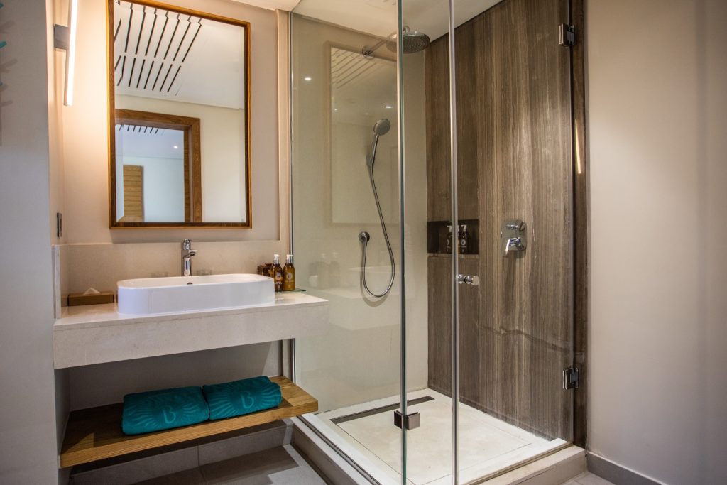 Modernes Badezimmer mit verglaster Dusche, Steinverkleidung, Holzregal und türkisfarbenen Handtüchern im Paradis Beachcomber.