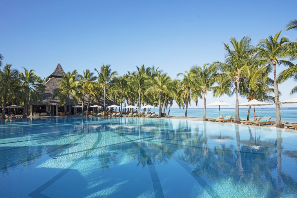Weitläufige Poollandschaft mit blau schimmerndem Wasser, Palmen, weißen Sonnenschirmen und Gebäude mit Strohdach