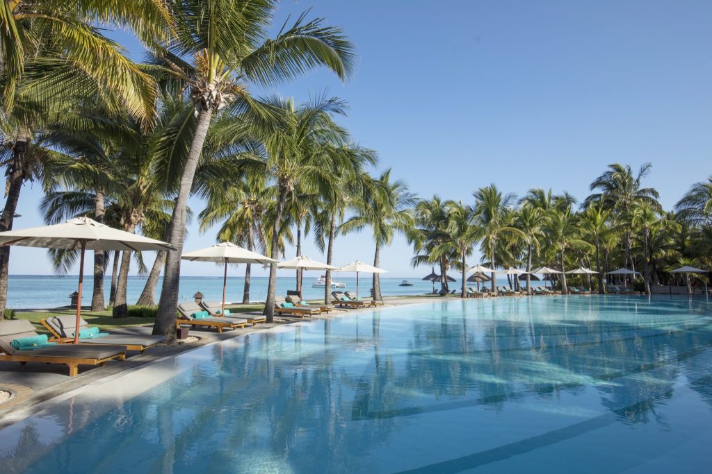 Großer Swimmingpool mit Holzliegen und hellen Stoffschirmen, Palmen und Meer im Hintergrund