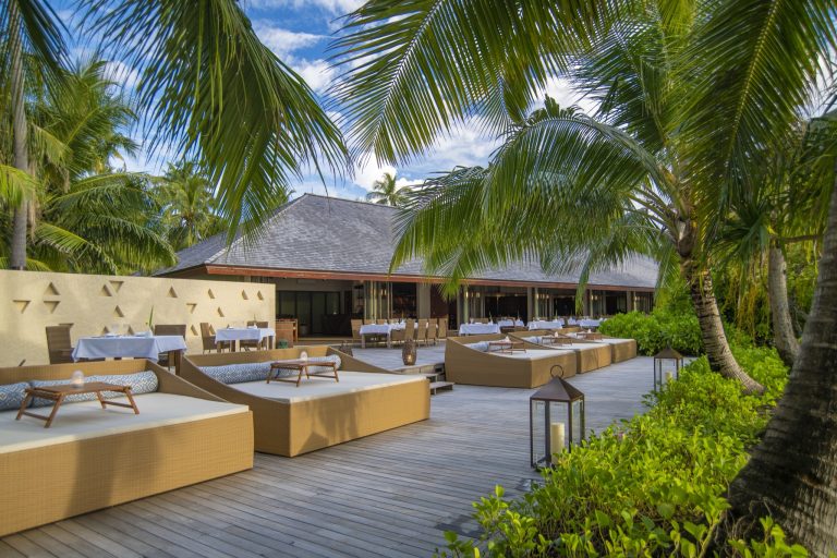 Terrasse mit beigefarbenen Rattanliegen, weißen Kissen, Holzdielen, Laternen und Restaurantbereich