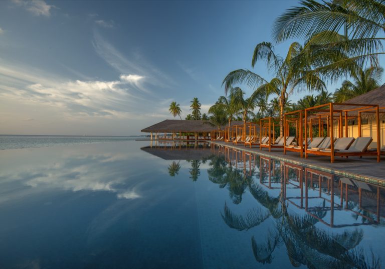 Infinity-Pool mit Holzliegen und Sonnendächern, Palmen spiegeln sich im Wasser