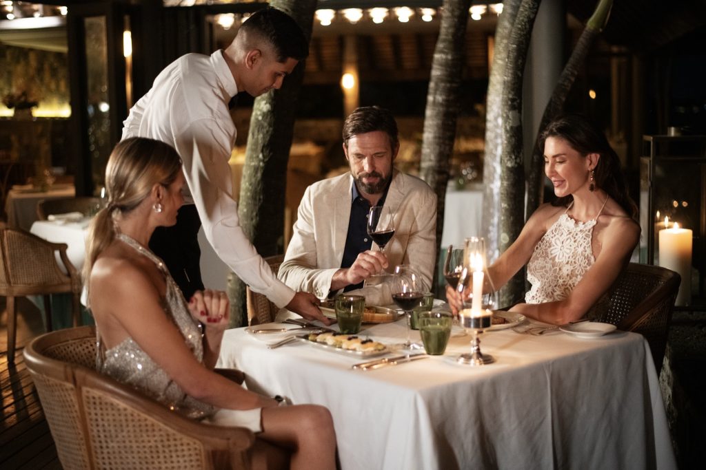 Tisch mit weißem Tischtuch und Kerzenlicht im Außenbereich eines Restaurants mit Holzboden und Bäumen im Hintergrund