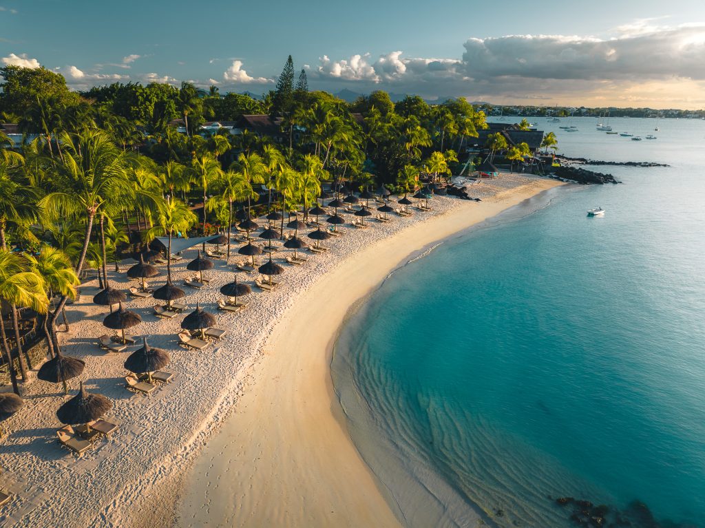 Strand mit gleichmäßig angeordneten Sonnenschirmen aus Naturmaterialien, grünen Palmen und türkisfarbenem Ozean bei Royal.