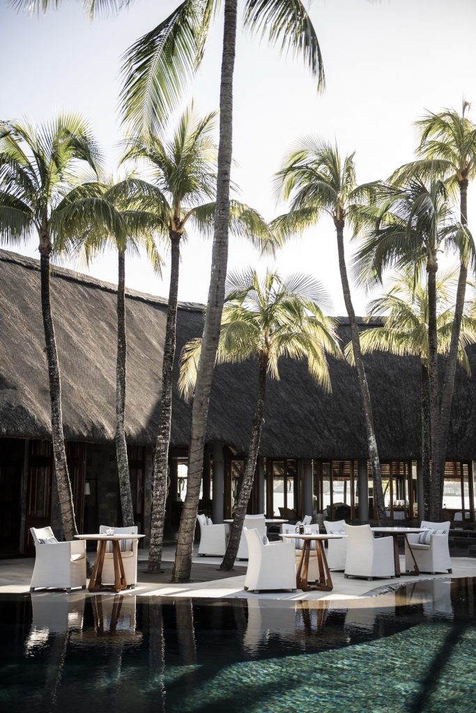 Außenanlage mit hohen Palmen, Reetdachgebäude, weißen Sesseln und Pool im Royal Palm Beachcomber Luxury