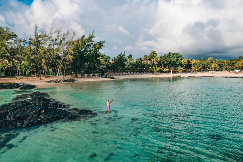 Strand mit türkisfarbenem Wasser, Palmen am Ufer, Fischer mit Angel neben dunklen Lavasteinen und Regenbogen im Hintergrund