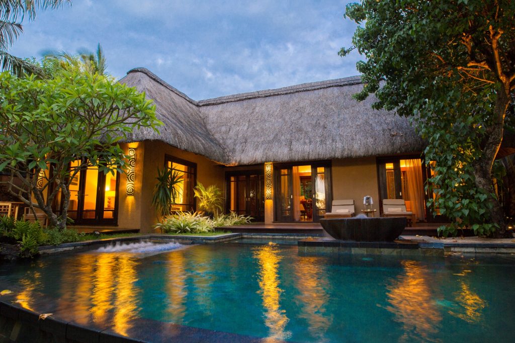 Hotelbungalow mit Strohdach, großflächigen Fenstern, Pool und üppiger Vegetation bei Dämmerung in Mauritius