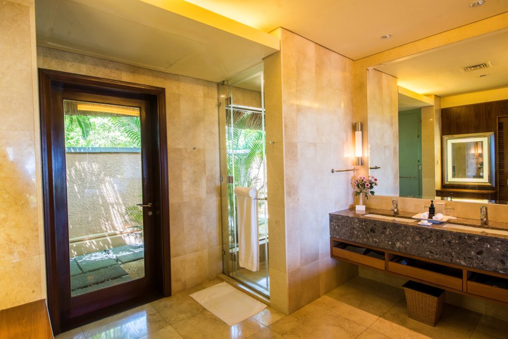 Badezimmer der Oceanview Pool Villa mit Holztür, zwei Waschbecken und cremefarbenem Steinboden