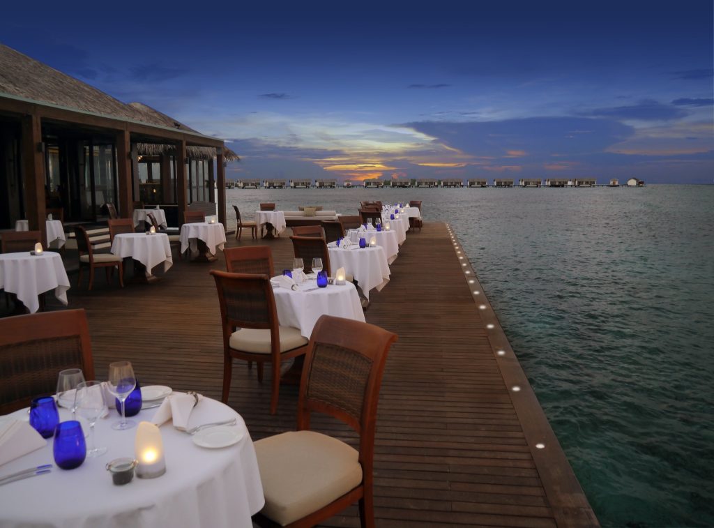 Terrasse des Restaurants The Falhumaa mit weißen Tischen, blauen Gläsern und Holzstühlen über dem Wasser bei Sonnenuntergang