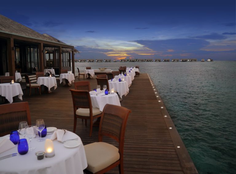 Terrasse des Restaurants The Falhumaa mit weißen Tischen, blauen Gläsern und Holzstühlen über dem Wasser bei Sonnenuntergang