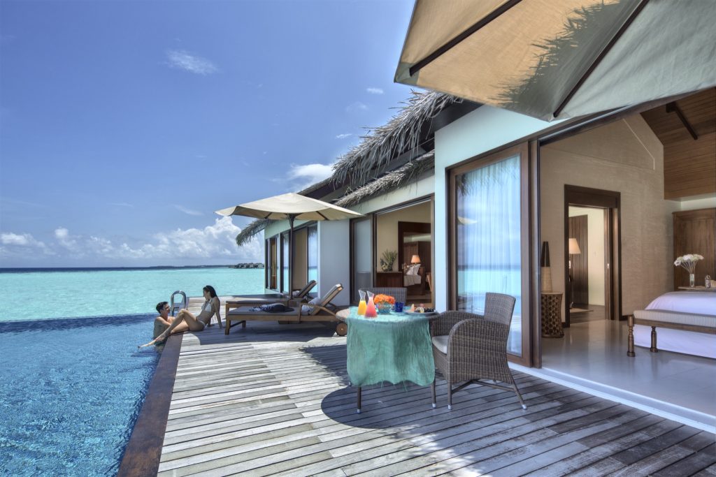 Terrasse mit Holzboden, türkisfarbener Tischdecke, Rattan-Stühlen, Poolliegen und Hotelzimmer mit großen Fenstern