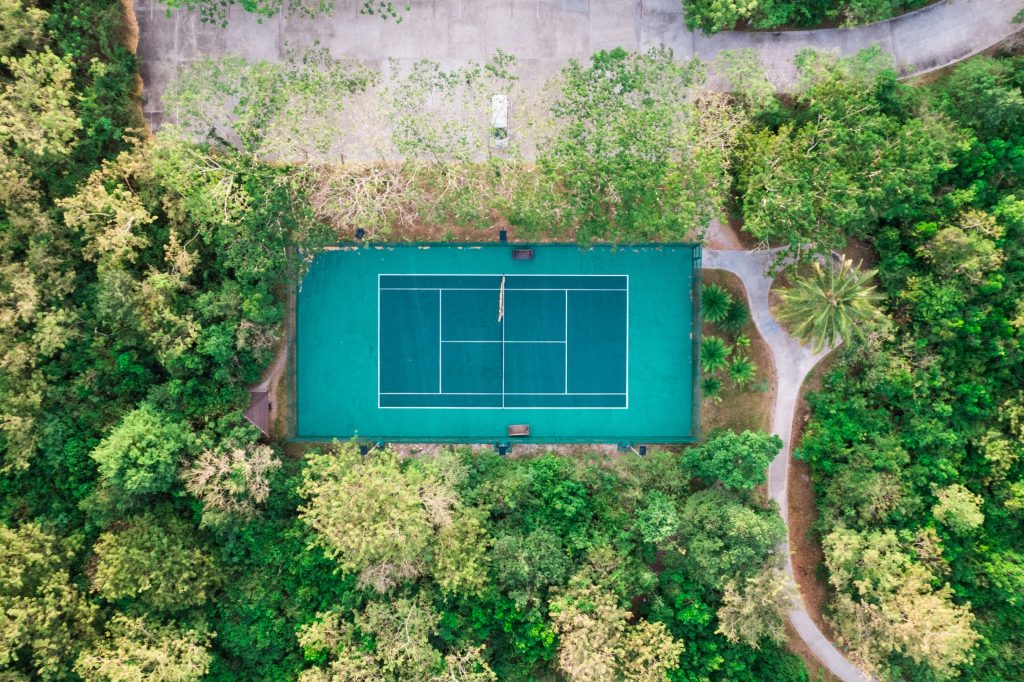 Vogelperspektive auf einen türkisfarbenen Tennisplatz mit weißen Linien umgeben von grünen Bäumen bei Tageslicht