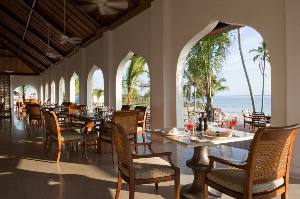 Restaurantterrasse mit weißen Bögen, Holztischen, Rattanstühlen, Palmen und Ozean im Hintergrund