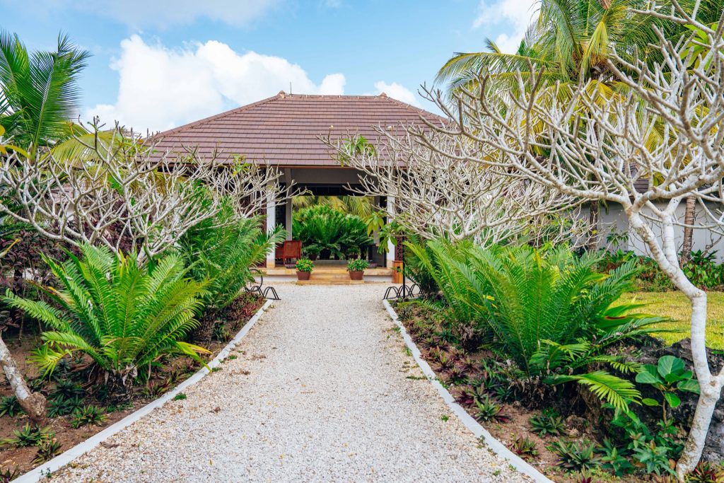 Weißer Kiesweg durch grünen Garten mit Farnen und kahlen Bäumen zum Wellness-Gebäude in The Residence Zanzibar