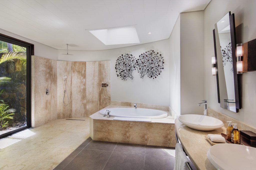 Modernes Badezimmer der Beachfront Suite mit freistehender Badewanne, Regenbrause und Fenster mit Blick auf tropische.