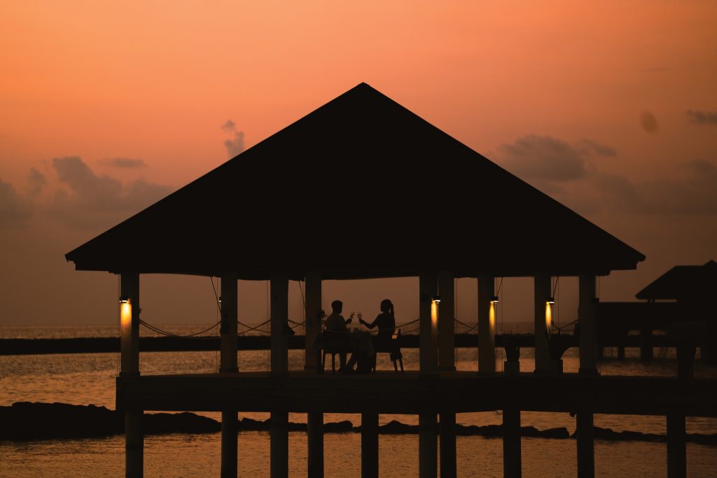 Pavillon mit Holzdach auf Pfählen im Wasser, zwei Personen an Tisch unter warmem Licht bei Sonnenuntergang