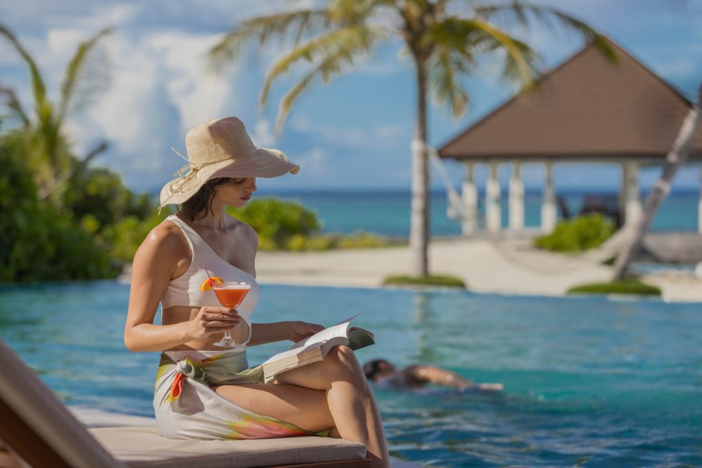 Frau mit Strohhut sitzt am Pool und liest ein Buch, hält Cocktailglas, Meer und Pavillon im Hintergrund