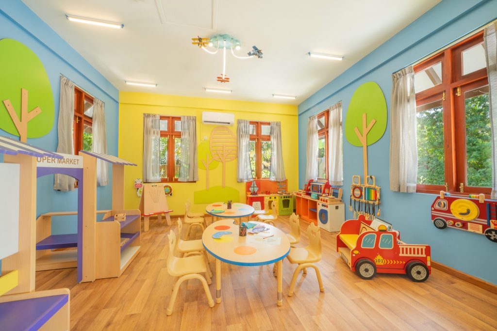 Bunter Kinderspielraum mit runden Holztischen, blauen und gelben Wänden, großen Fenstern und Spielstationen im Varu by.