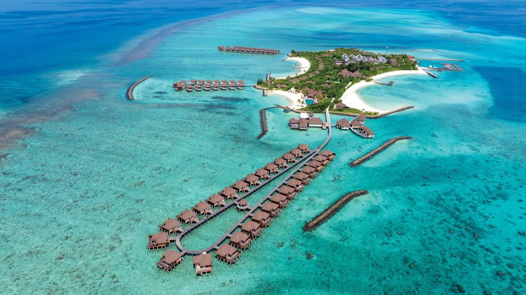Vogelperspektive auf Wasserbungalows auf Stelzen über klarem türkisfarbenem Meer mit Holzstegen zur Insel