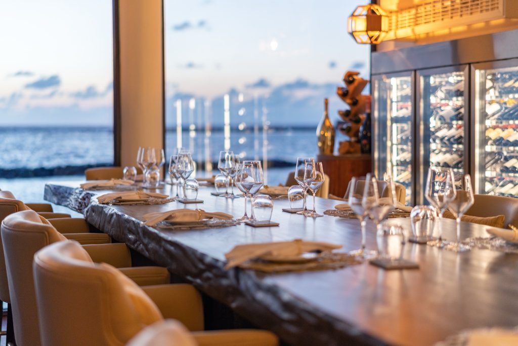 Tisch aus dunklem Holz mit Gläsern, Tellern und Stoffservietten im Restaurant Cellar at Nu mit Weinregalen und Meerblick