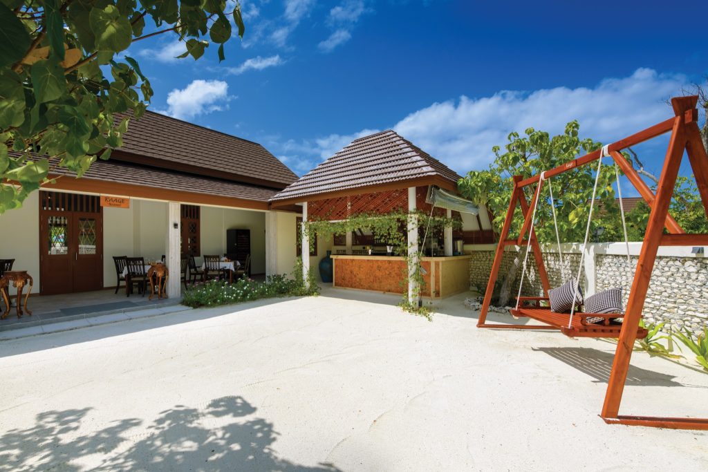 Offene Hotelarchitektur mit Holzpavillon, Sandboden, Tisch mit Stühlen links und Schaukel rechts im Restaurant Kaage