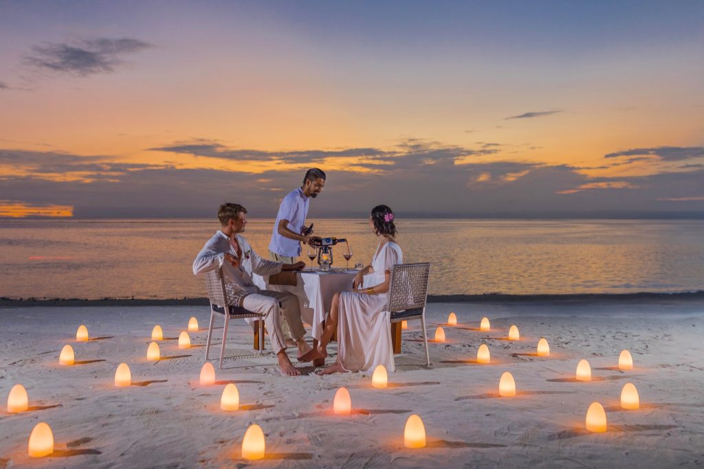 Paar sitzt an einem Tisch mit weißem Tischtuch und Laternen am Strand bei Sonnenuntergang im Varu by Atmosphere Restaurant