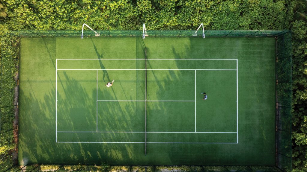 Tennisplatz mit grünem Bodenbelag und weißen Linien, umgeben von dichtem Grün, zwei Spieler in Aktion
