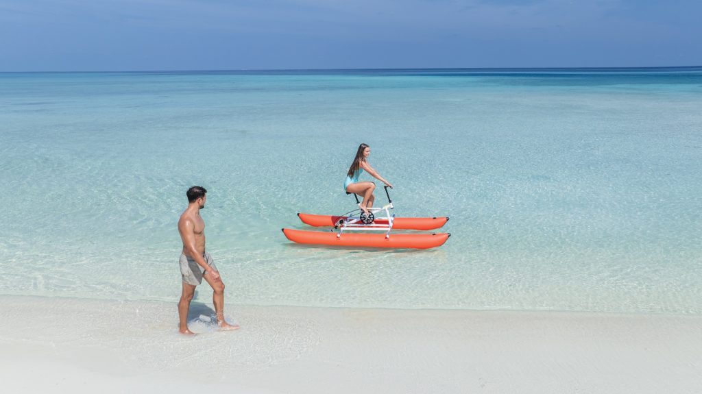Frau auf Wasserfahrrad mit orangen Pontons im klaren Wasser, Mann am Wasserrand, Strand mit weißem Sand